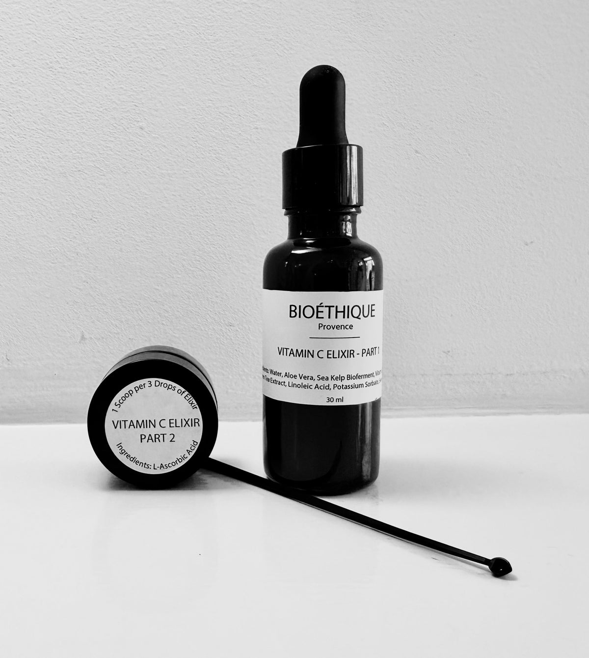 A black bottle with a dropper labeled 'VITAMIN C ELIXIR - PART 1' next to a jar labeled 'VITAMIN C POWDER - PART 2' with a spoon inside the jar. The bottle and jar are placed on a white surface.