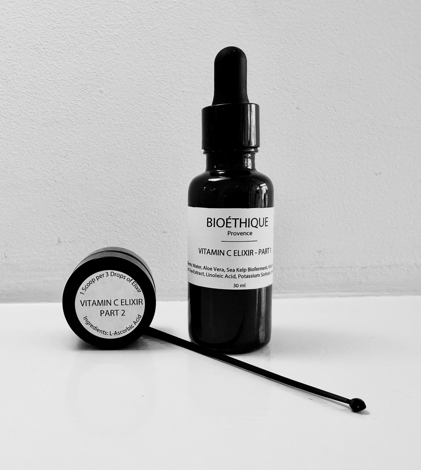 A black bottle with a dropper labeled 'VITAMIN C ELIXIR - PART 1' next to a jar labeled 'VITAMIN C POWDER - PART 2' with a spoon inside the jar. The bottle and jar are placed on a white surface.